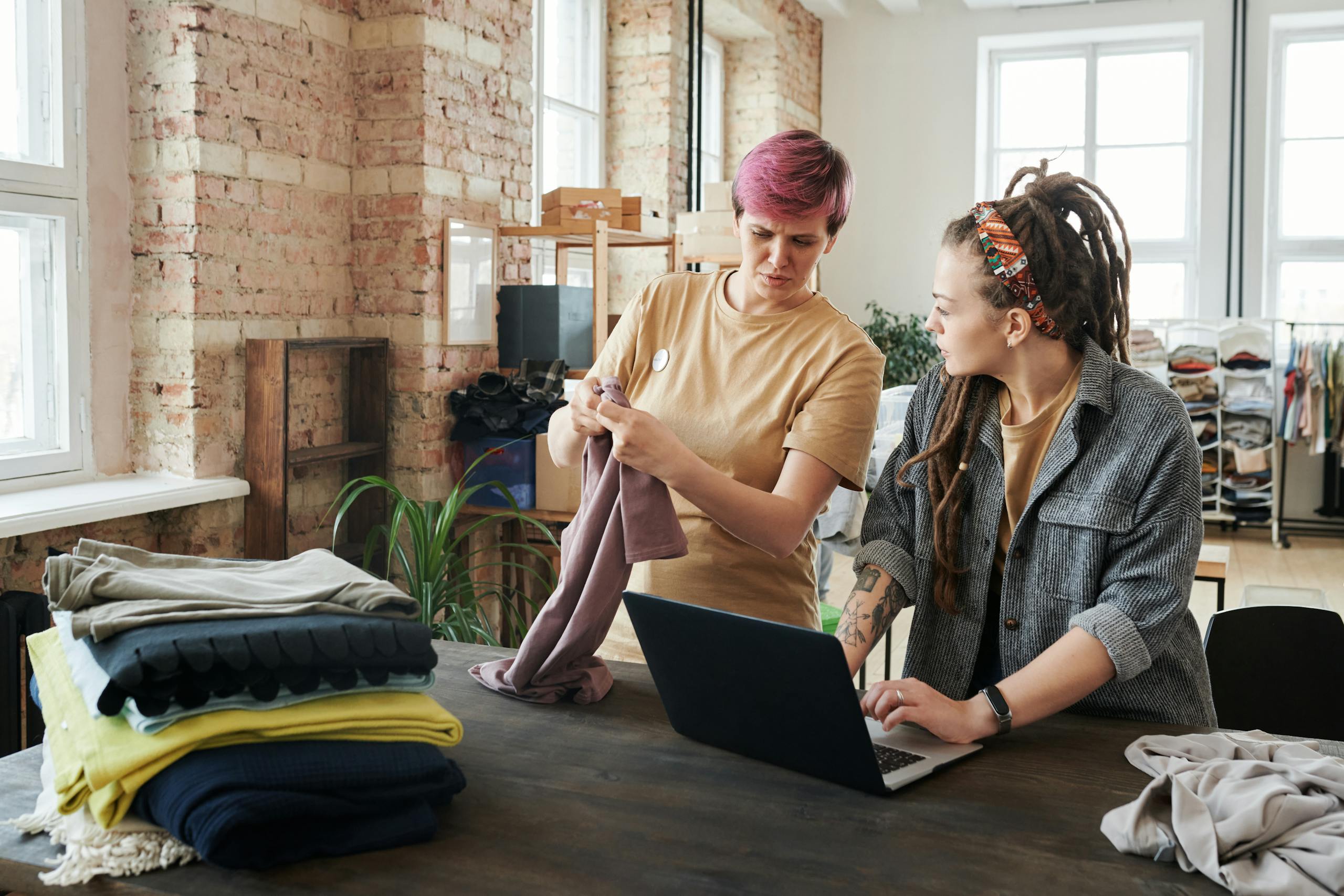 "doing good with technology" Two women sorting clothing donations using a laptop in a bright urban setting.